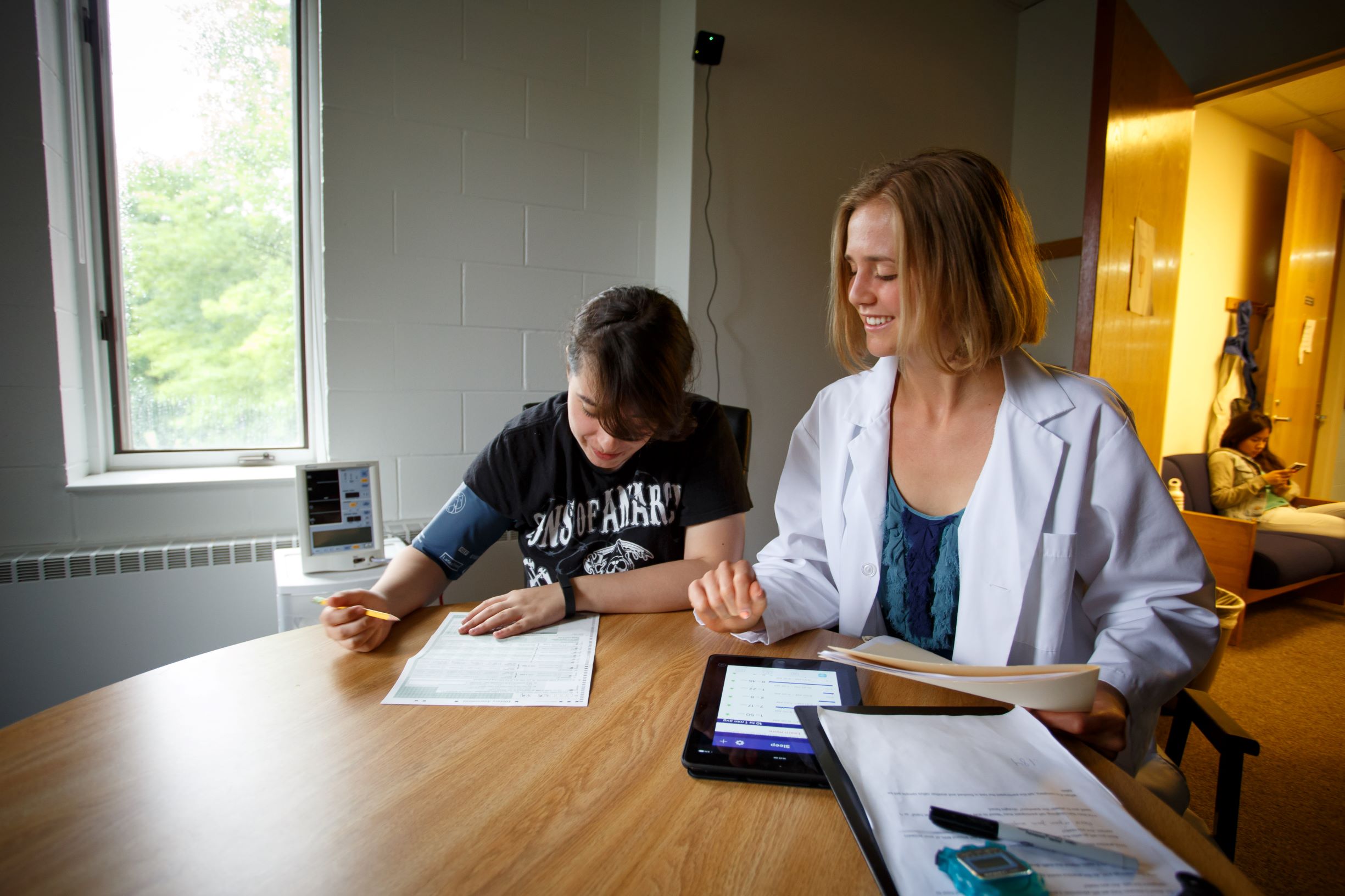 student wearing a lab coat stands next to a student writing at a table.