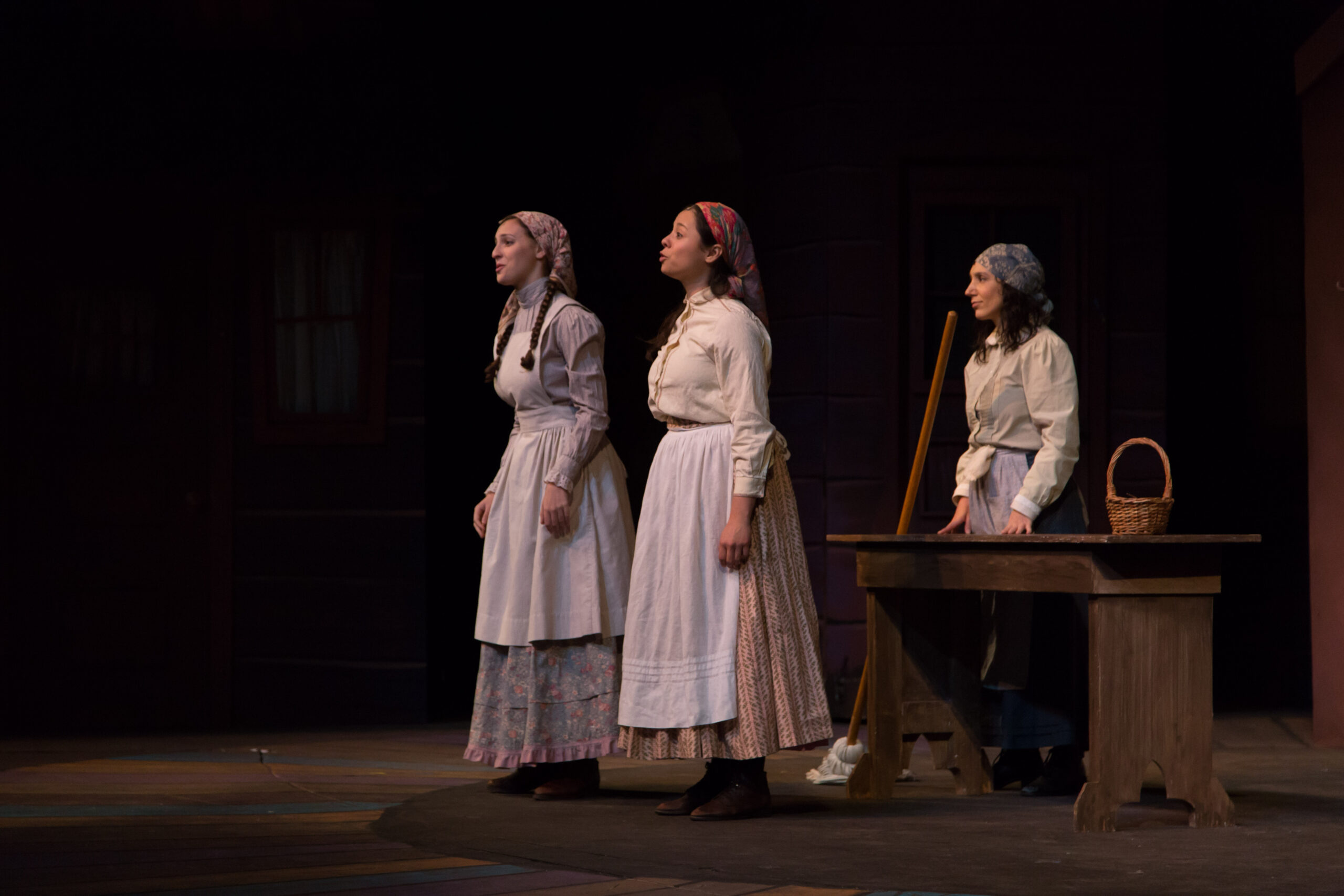 thre women in profile sing on a darkened stage for a musical theatre production.